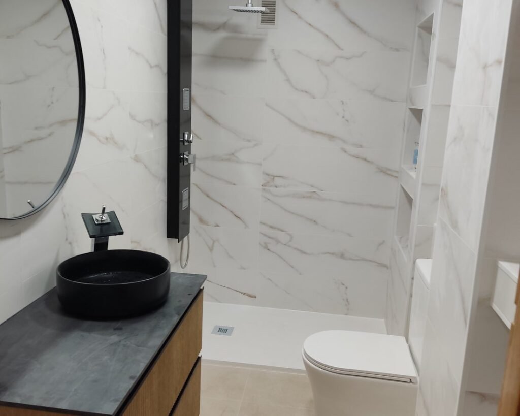 Baño reformado con azulejo tipo mármol, lavabo negro sobre encimera y columna de ducha