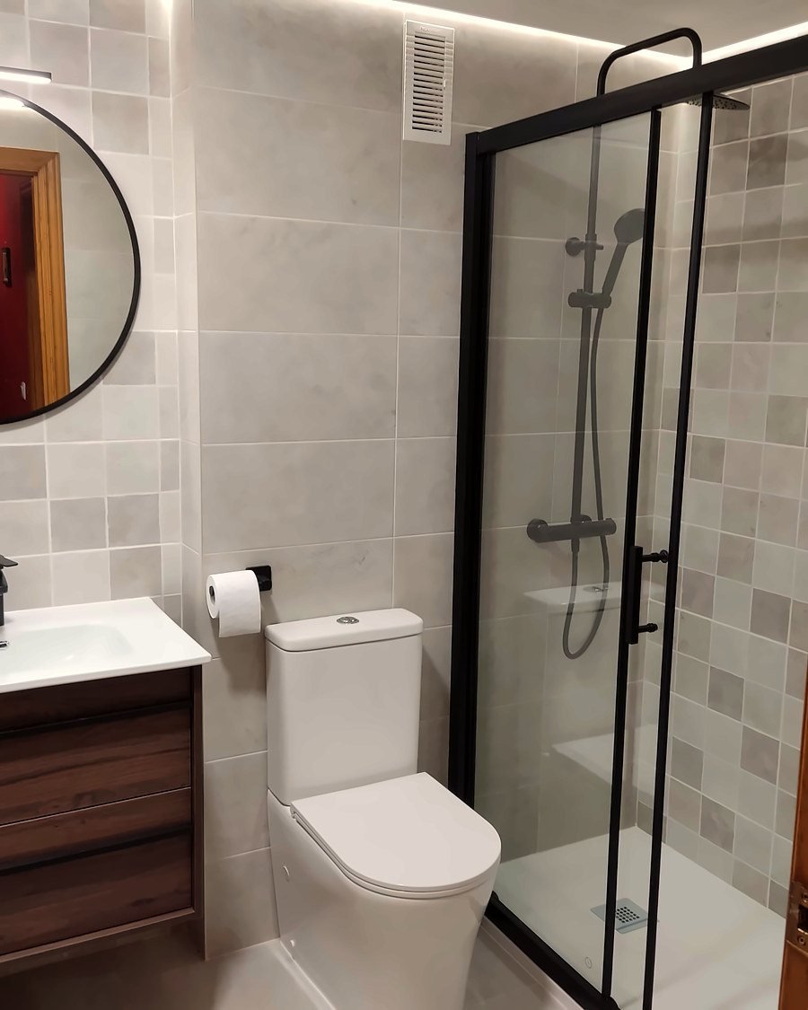 El mismo baño reformado, con plato de ducha, mampara negra y mueble de madera